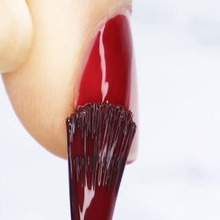 A close-up of a nail being painted with a glossy dark red nail polish. The brush is applying the polish smoothly.